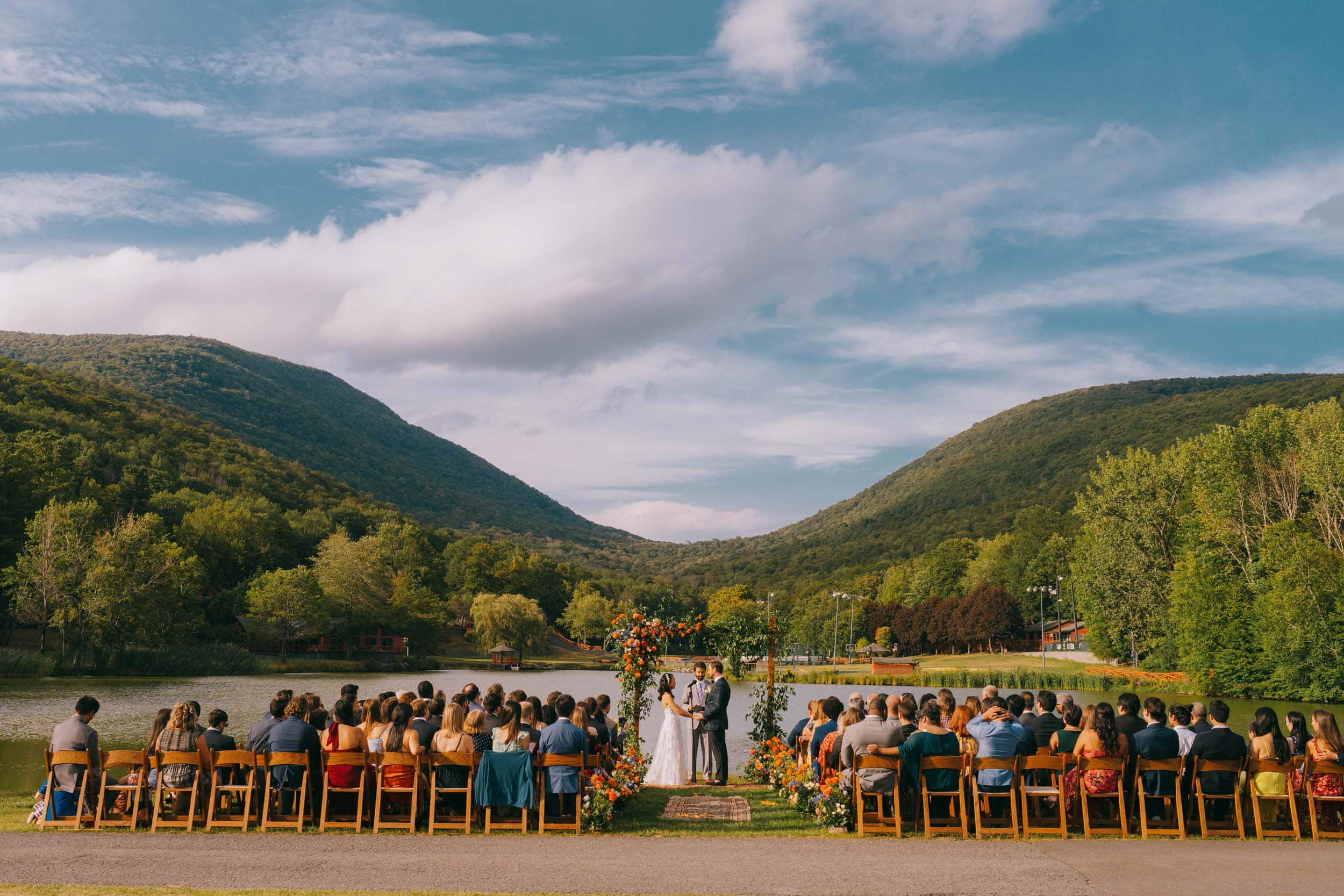 Outdoor Hudson Valley wedding ceremony with lake and mountains in the background and wooden chairs with guests
